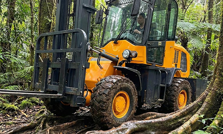A rough terrain forklift with high ground clearance driving over a series of large, exposed tree roots.